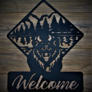 metal sign of a bear standing in front of a forest that is framed in a diamond shape with the word Welcome in script below the bear