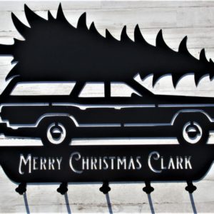 A black metal sign of a station wagon transporting a Christmas tree with the words Merry Christmas Clark below the wheels along with five coat hooks