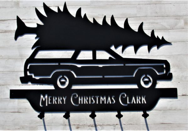 A black metal sign of a station wagon transporting a Christmas tree with the words Merry Christmas Clark below the wheels along with five coat hooks